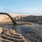 Well water sediment build-up. Pipe discharging dark, sediment-filled water into a sandy area. Water testing for well owners.