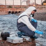 PFAS in well water: Technician in protective suit collecting water sample for PFAS testing near industrial runoff.