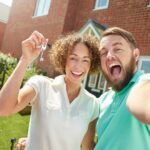 Happy new home buyers holding keys in front of new house. Buying property tips.